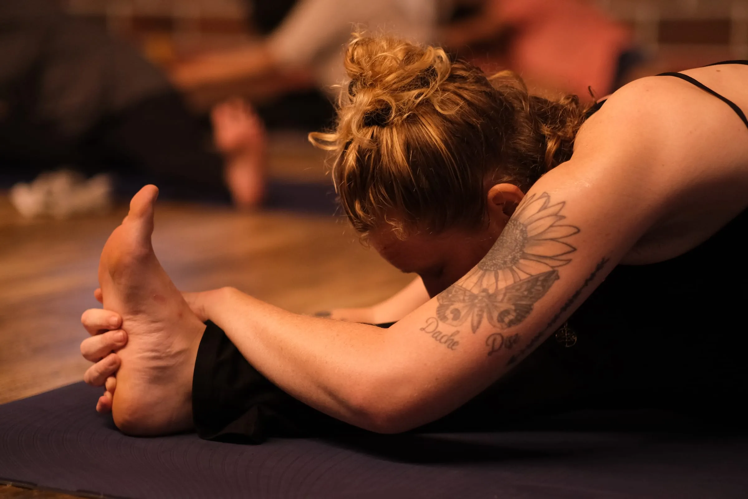 Laura en flexion avant sur sa jambe tendue, tenant son pied, dans un studio à la lumière chaude