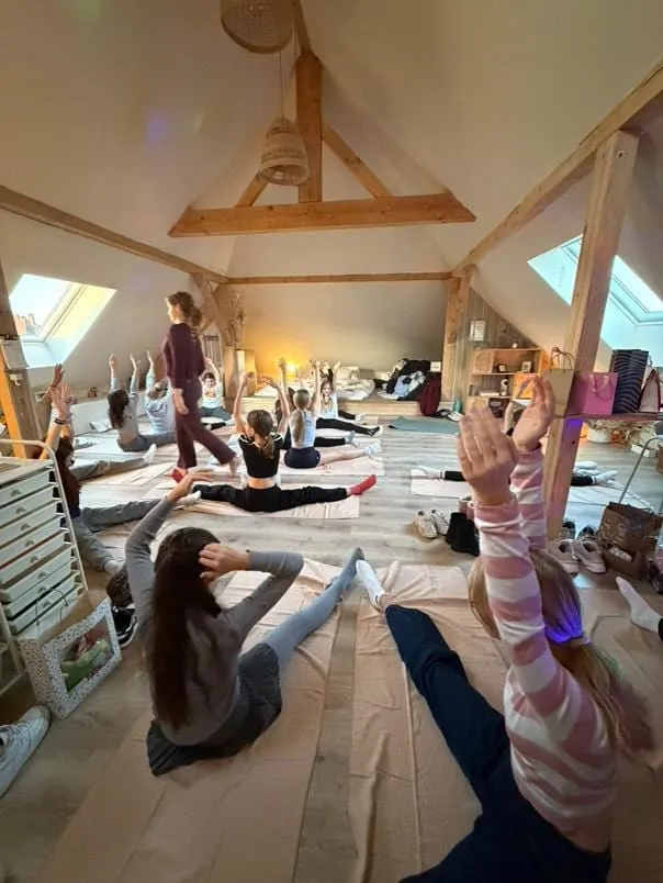 Un groupe pratique le yoga dans une pièce mansardée en bois, Laura passe entre les tapis